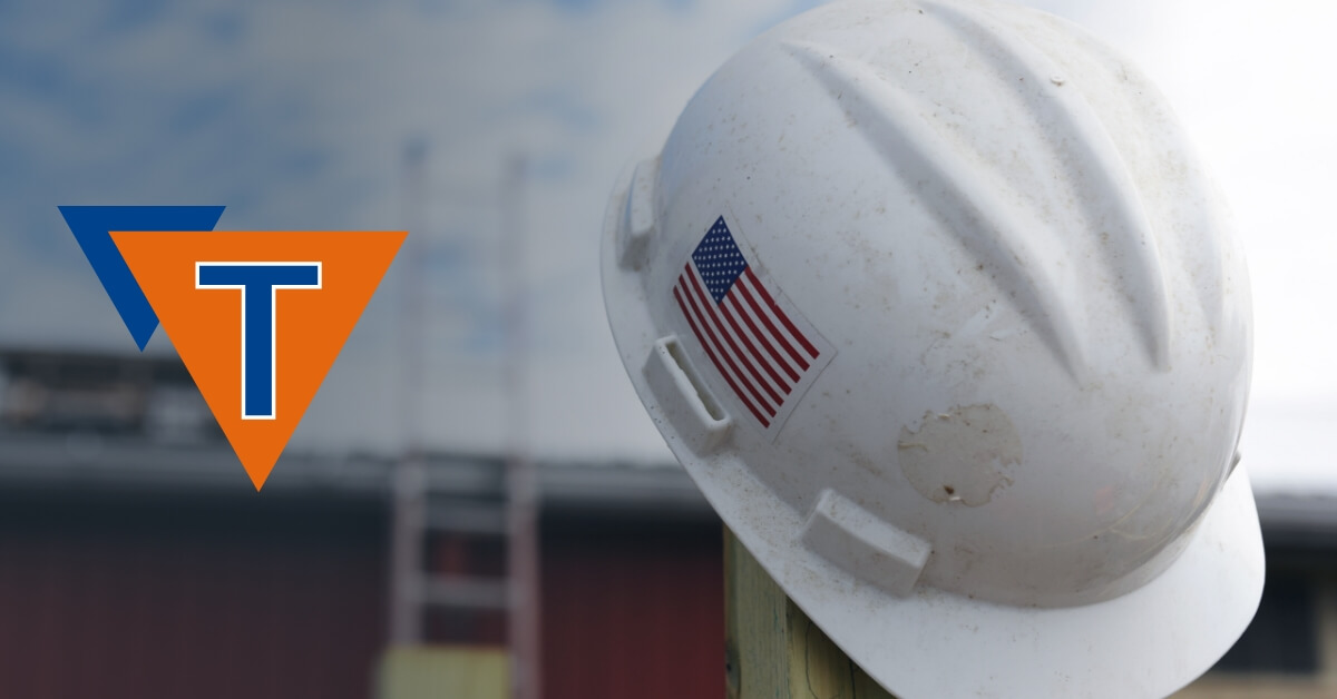White hard hat with an American flag sticker on a blurred background with the Triangle Manufacturing Logo.
